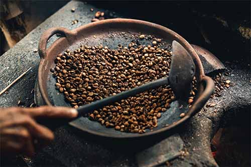 A man roasting coffee beans in an iron skillet. The different variety of coffee beans impact flavour, this blog on Bean Merchant's website talks about how.
