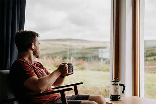 A man sits with a plunger or French press and a cup of coffee in his hand staring at the scenery. This image is at the top of Bean Merchant's guide to making the best plunger coffee in the world.